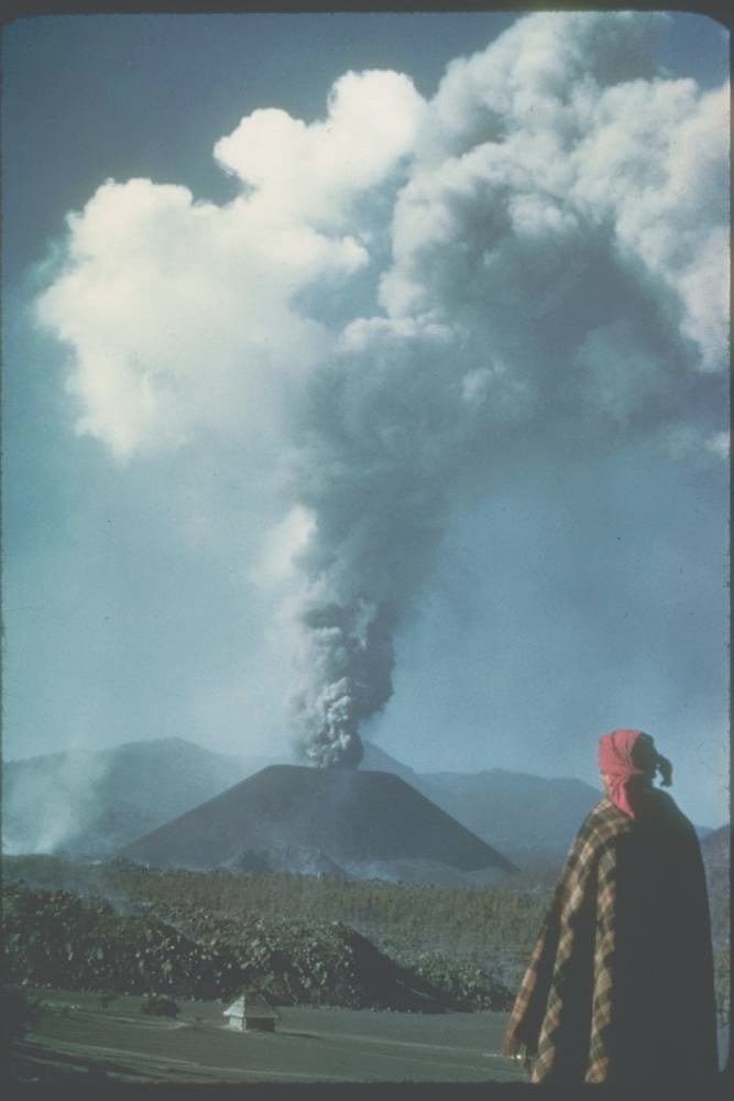 Parícutin en erupción, con una columna elevada, el cono, campos y una persona en primer término.